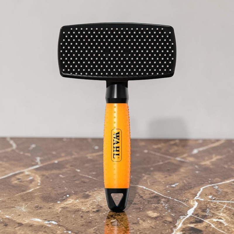 Große, rechteckige Slicker-Bürste mit schwarzem Kopf und vielen hellen Nylonborsten. Ergonomischer, leuchtend orangefarbener, texturierter Griff mit WAHL-Logo. Unten schwarze Aufhängeschlaufe. Auf brauner, marmorierter Oberfläche.