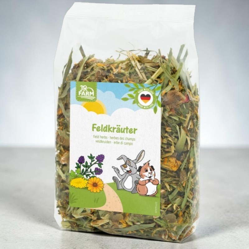 Transparente Tüte mit getrockneten Feldkräutern für Heimtiere; sichtbar Gräser, Blätter und Blüten. Etikett: JR FARM Logo, Produktname „Feldkräuter“ und Illustration eines lachenden Hasen und Meerschweinchens auf Blumenwiese.