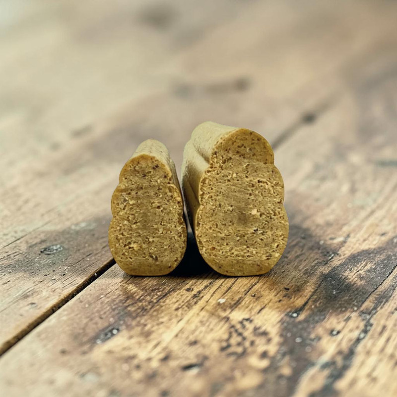 Zwei aufrecht stehende, längliche Hundesnacks mit unregelmäßiger, leicht gewellter Form auf rustikaler Holzoberfläche. Hellbraun, feinkörnig, mit kleinen dunkleren Einschlüssen. Links schmaler als rechts.