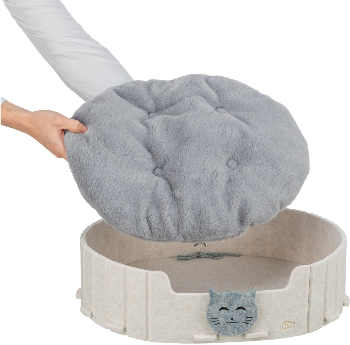 Le mani inseriscono un morbido cuscino rotondo in peluche grigio in una cuccia rotonda color crema in feltro con un allegro gattino ricamato sul davanti. Il cuscino trapuntato con bottoni si adatta perfettamente e invita al relax.