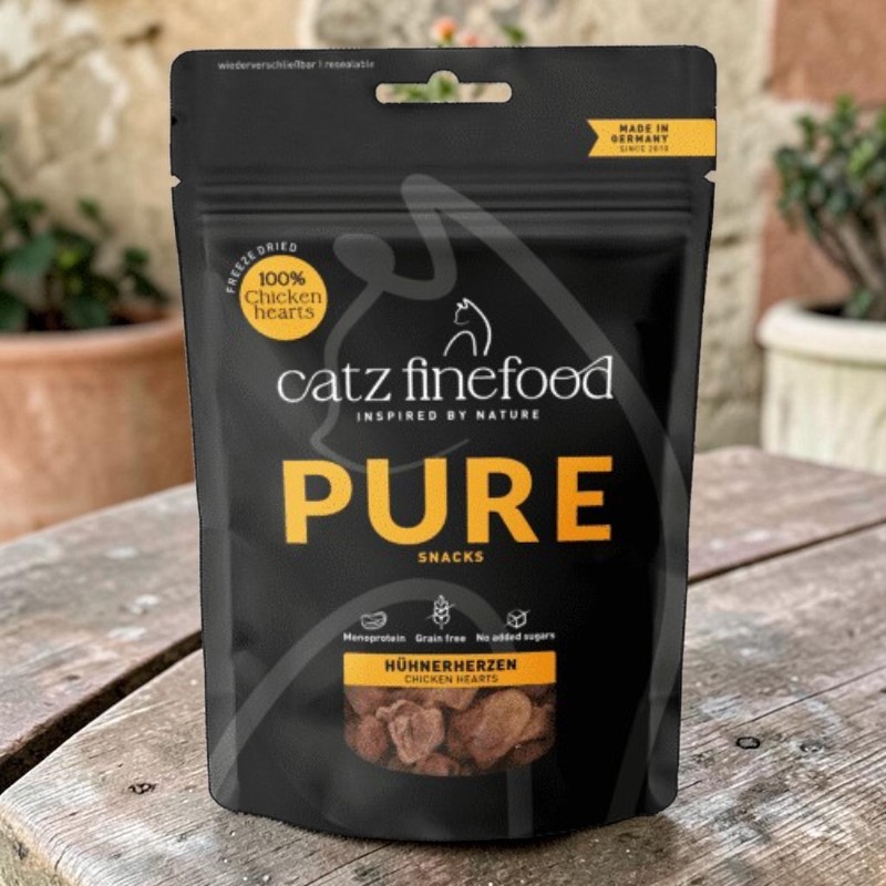 Confezione di snack Catz Finefood PURE nera (100% cuori di pollo liofilizzati). Su tavolo di legno, scritta PURE in oro. Note: monoproteico, senza cereali, senza zuccheri aggiunti.