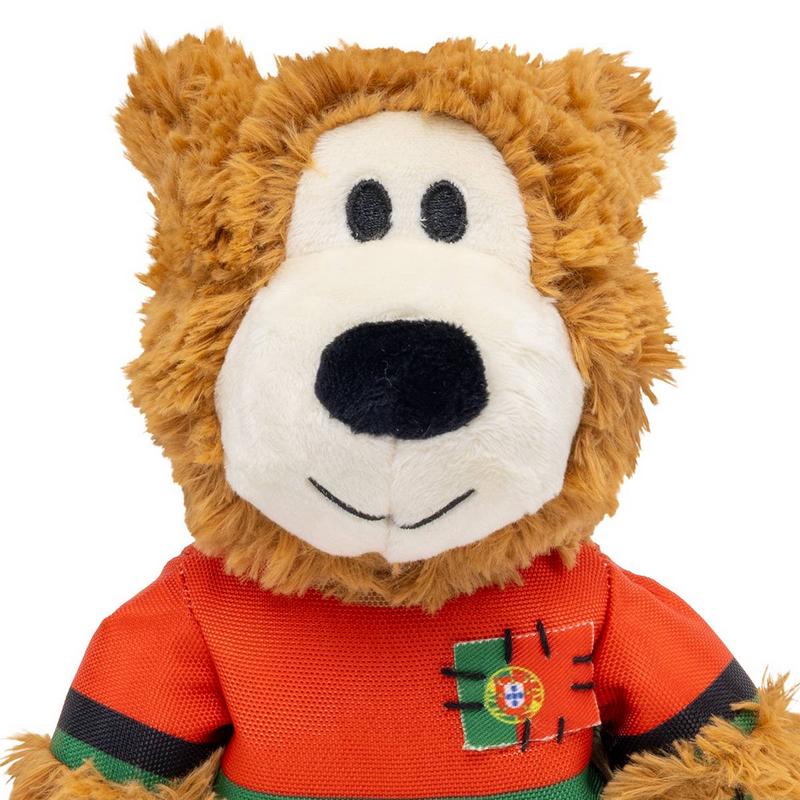 Testa in peluche marrone, morbida, con viso bianco, occhi a bottone neri, grande naso nero e un accenno di sorriso. Indossa una maglietta rossa con strisce nere e verdi sulle maniche e una toppa con la bandiera portoghese sul petto.