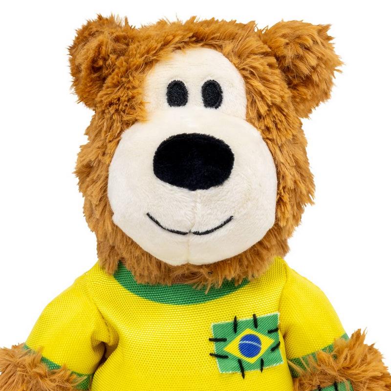 Un orsacchiotto di peluche marrone dal pelo folto, con il muso chiaro e il naso nero, guarda dritto verso la fotocamera. Indossa una maglietta gialla con strisce verdi, con lo stemma della bandiera brasiliana.