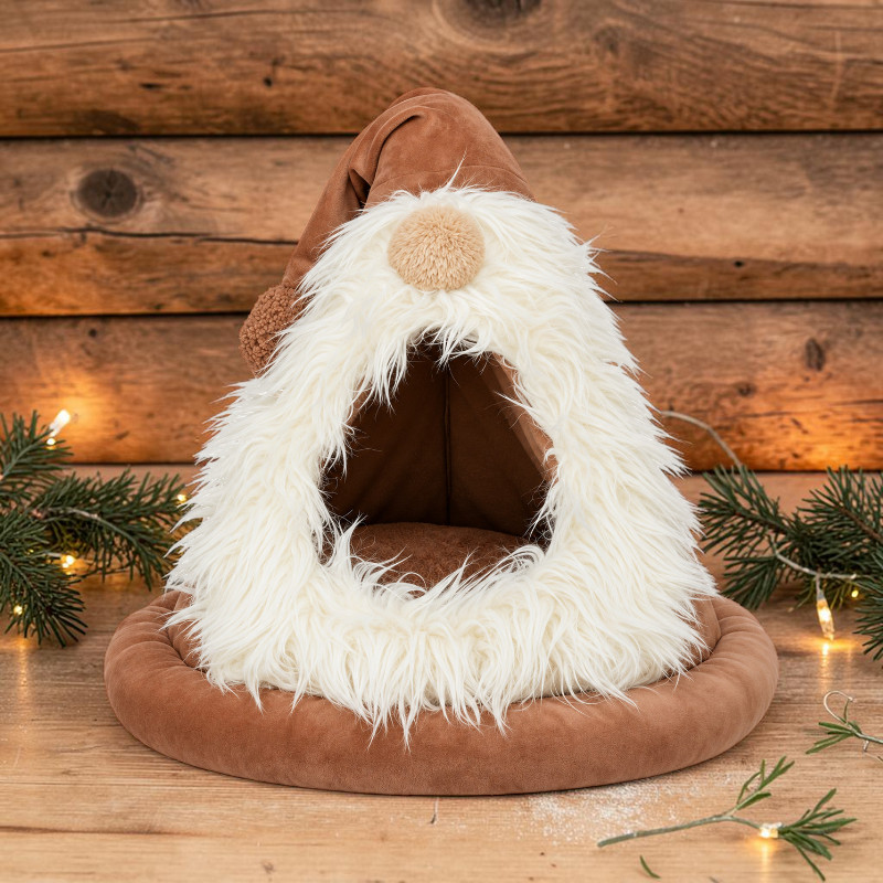 Una cuccia per animali domestici marrone e morbida a forma di gnomo, con un cappellino a punta e una soffice barba bianca che funge da ingresso. Si trova su un pavimento in legno con decorazioni natalizie come rami di abete e lucine sullo sfondo.