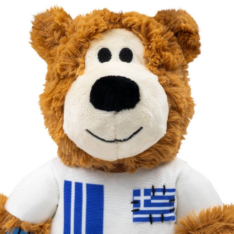 Ein flauschiger, brauner Teddybär schaut nach vorne. Er hat eine cremefarbene Schnauze, runde schwarze Augen, eine schwarze Nase und ein gesticktes Lächeln. Er trägt ein weißes Hemd mit zwei blauen Streifen und einer kleinen griechischen Flagge.