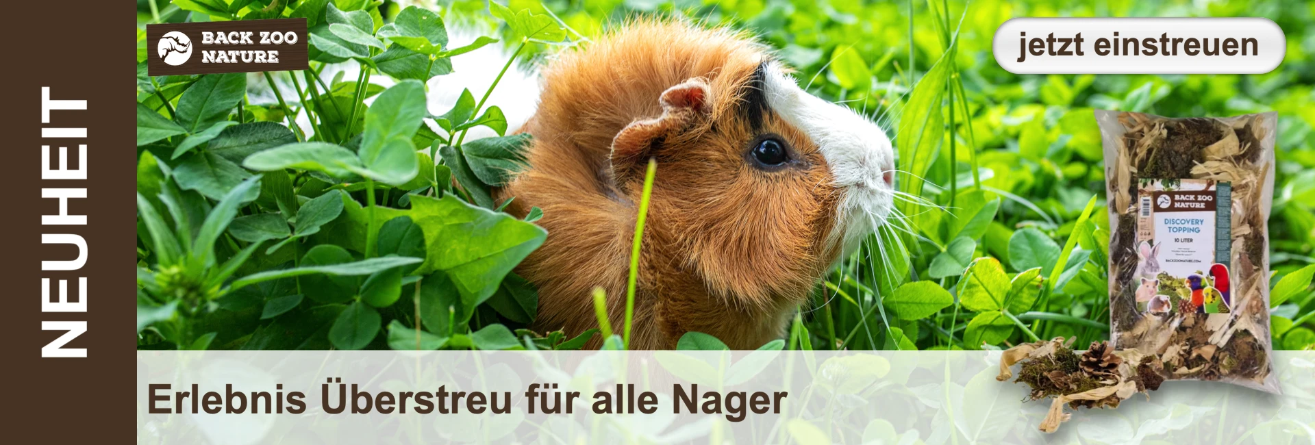 Ein braun-weißes Meerschweinchen sitzt im grünen Klee; rechts davon eine Verpackung "Discovery Topping" für Nager.