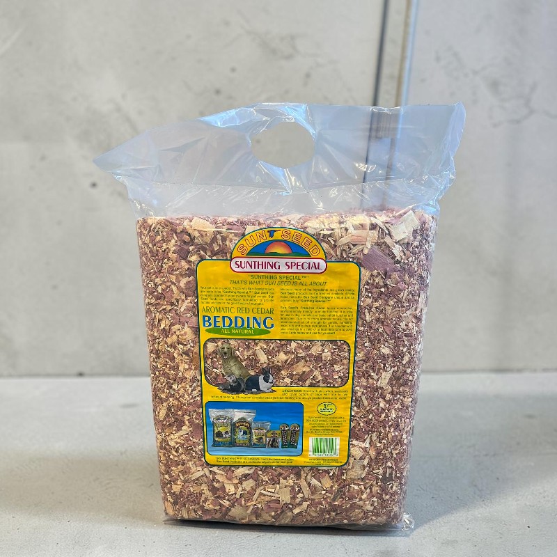 Grande sacco di plastica trasparente con lettiera aromatica di legno di cedro bruno-rossastro. Etichetta gialla: "AROMATIC RED CEDAR BEDDING ALL NATURAL" con immagini di animali. Su sfondo chiaro, davanti a parete di cemento grigio.