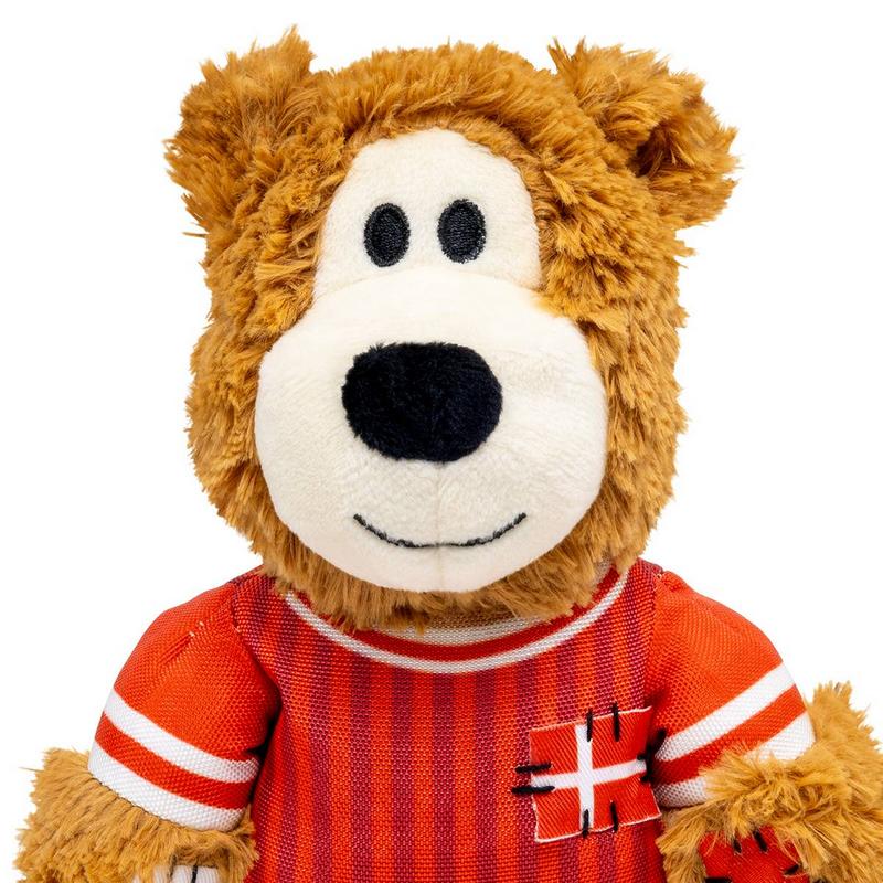 Orsacchiotto di peluche marrone con pelo soffice dalla testa in su. Indossa una maglietta a righe rosse e bianche con una piccola toppa con la bandiera danese cucita sul lato destro del petto. Faccia bianca, occhi rotondi, naso e sorriso.