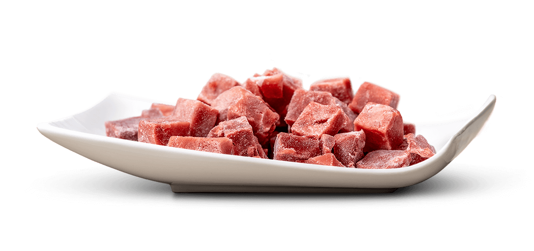 Weiße, geschwungene Schale mit vielen tiefgefrorenen, rotbraunen Fleischwürfeln. Unregelmäßig geschnitten, mit leicht rauer, frostiger Oberfläche und sichtbaren Eiskristallen, dicht beieinander angeordnet.