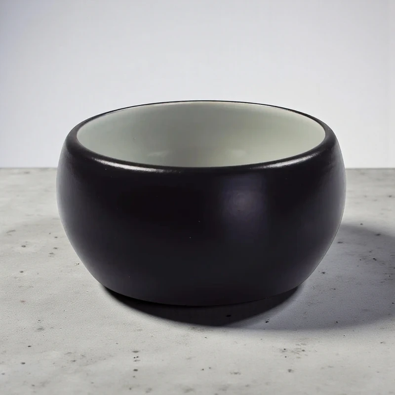 Black, round ceramic dog bowl with white inside, standing on a grey surface.