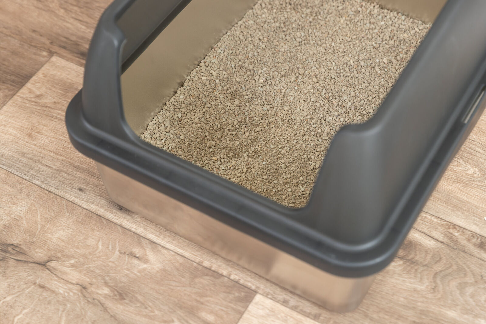 A clean cat litter tray with a shiny stainless-steel bowl and a dark, modern plastic frame is filled with light brown, grainy cat litter. It stands on a light-coloured wooden floor with a distinct grain. The view is from an angle above.