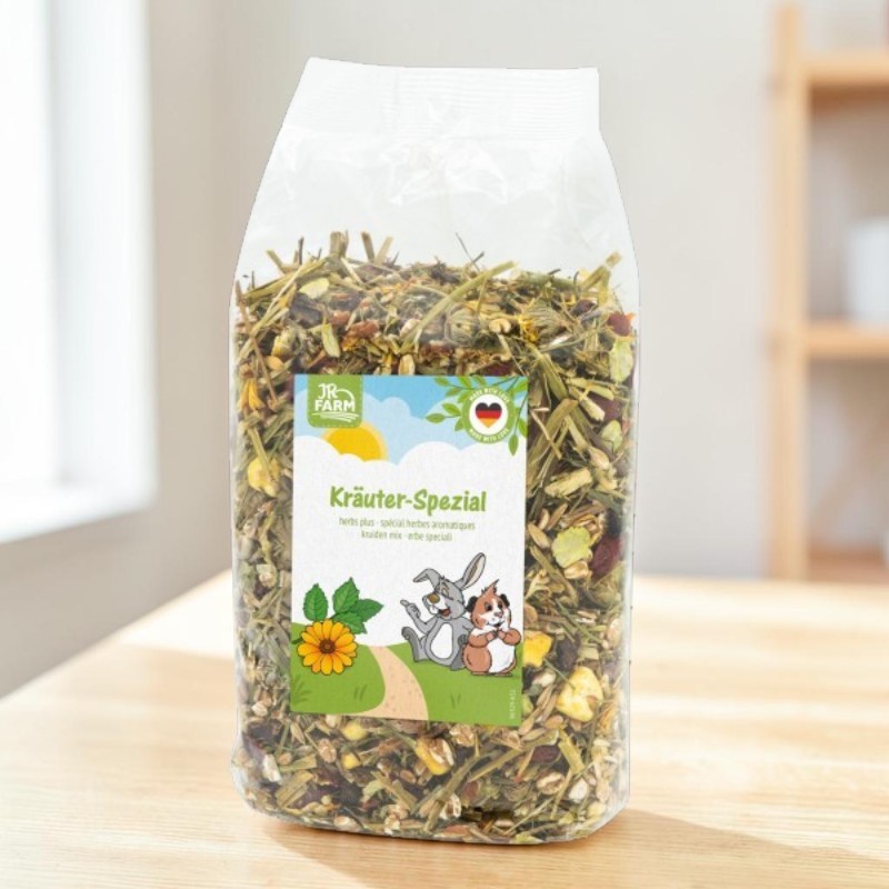 Transparent bag with JR Farm Herb Special: a mixture of dried herbs, hay and plant pieces for small animals. Label featuring a rabbit, a guinea pig, a yellow flower and the words ‘Herb Special’. On a light-coloured wooden table.