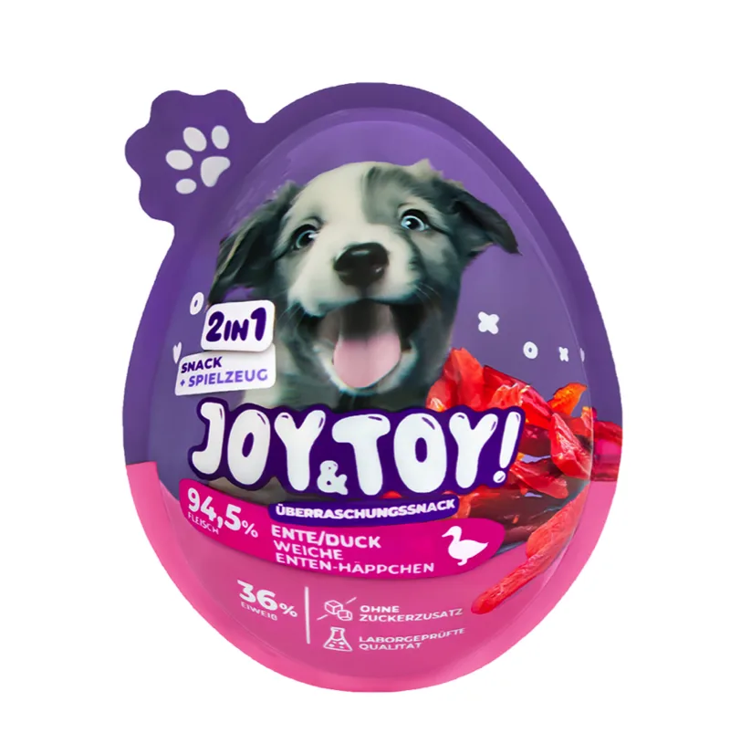 Lila und pinke Hundesnack-Verpackung in Eiform mit einem glücklichen Welpenbild. Beschriftungen zeigen "JOY & TOY!", "2IN1 SNACK + SPIELZEUG" und "94,5% FLEISCH", "ENTE/DUCK WEICHE ENTEN-HÄPPCHEN". Rechts sind rote, glänzende Entenhäppchen sichtbar.