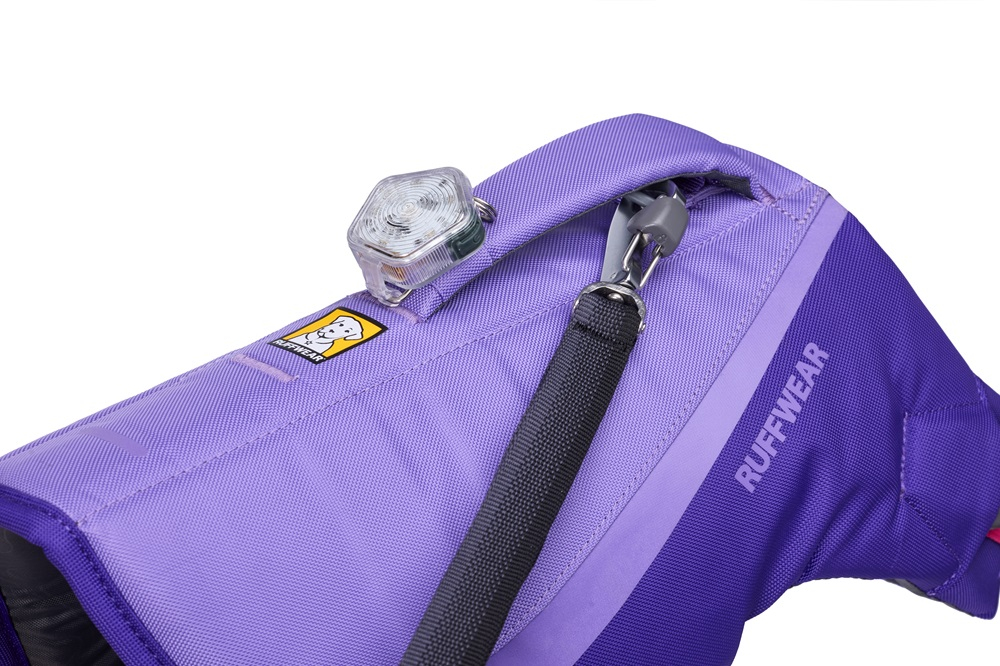 Gros plan sur un harnais pour chien « Ruffwear » violet, avec logo d'une tête de chien jaune. En haut, un gyrophare de sécurité carré transparent est fixé, ainsi qu'une laisse noire avec mousqueton. « Ruffwear » sur le côté.