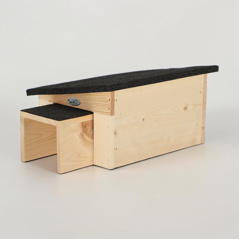 Maisonnette pour hérisson allongée, en bois d'épicéa clair, avec toit en pente recouvert de bitume gris foncé. À l'avant, petite entrée en saillie avec sa propre couverture en bitume. Grain du bois visible.