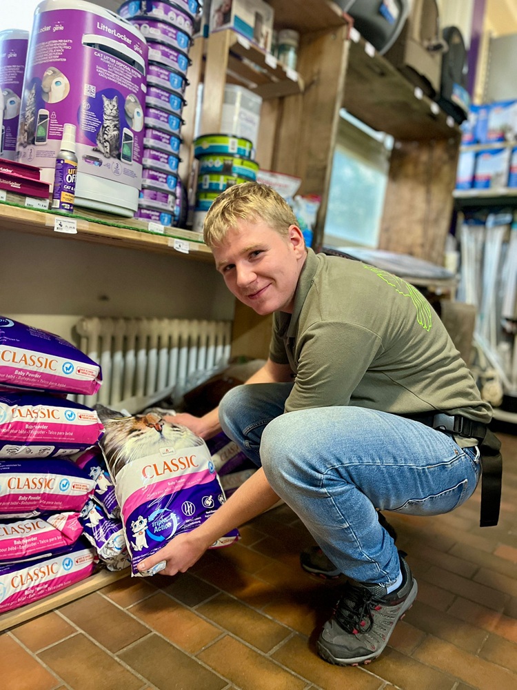 A man squats in front of a shelf of cat food and holds a packet of "Classic Baby Powder" in his hands.