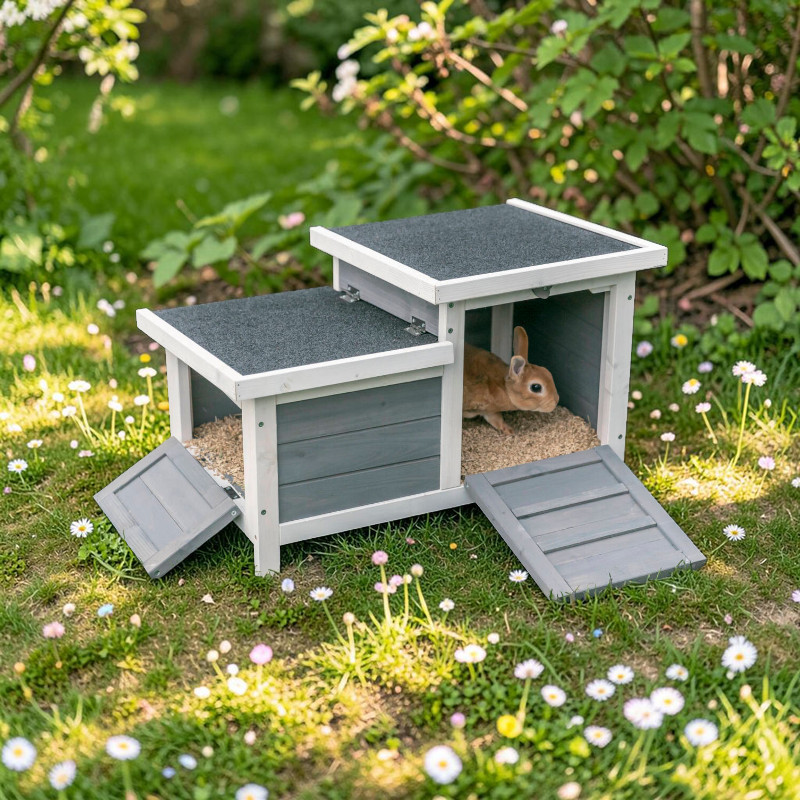 Zweistöckiges Kleintierhaus in Weiß und Grau auf grüner Wiese mit Gänseblümchen. Ein hellbraunes Kaninchen schaut unten aus Hobelspänen. Beide Eingänge mit dunkelgrauen Rampen offen. Dach mit Dachpappe. Büsche und Bäume im Hintergrund.
