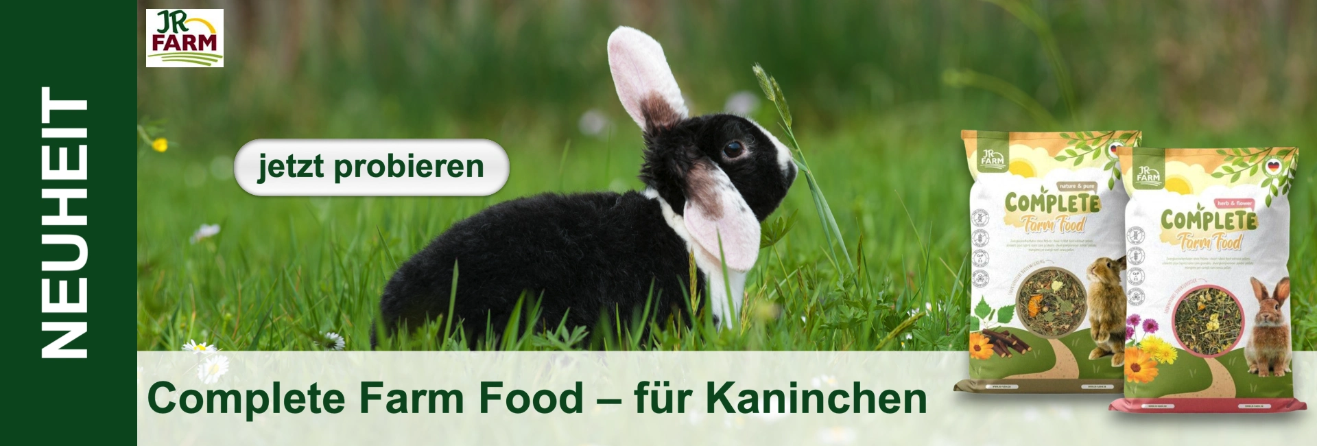 Flauschiges schwarz-weißes Kaninchen mit großen Ohren in saftig grüner Wiese mit kleinen weißen und gelben Blumen. Daneben zwei bunte Packungen JR FARM Complete Farm Food für Kaninchen. Oben links JR FARM Logo, „NEUHEIT“, Button „jetzt probieren“.
