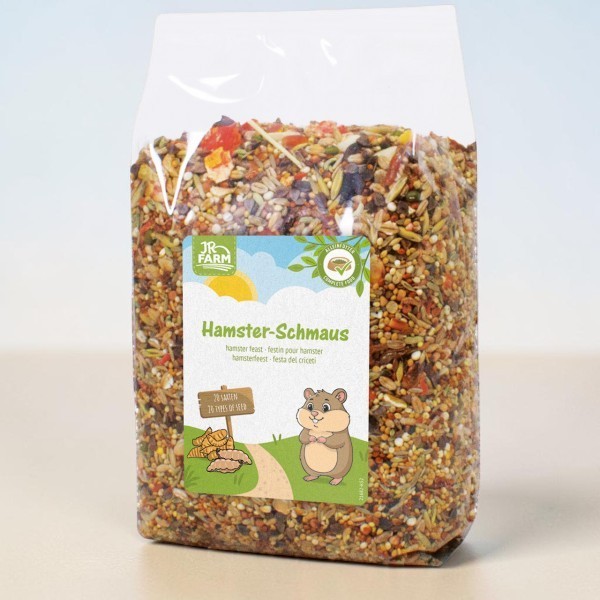 Transparent packaging containing JR Farm Hamster Treat, filled with a colourful mix of seeds, grains, dried fruit and vegetables. Label: a smiling cartoon hamster and a note stating that the meal contains 20 different types of seeds.