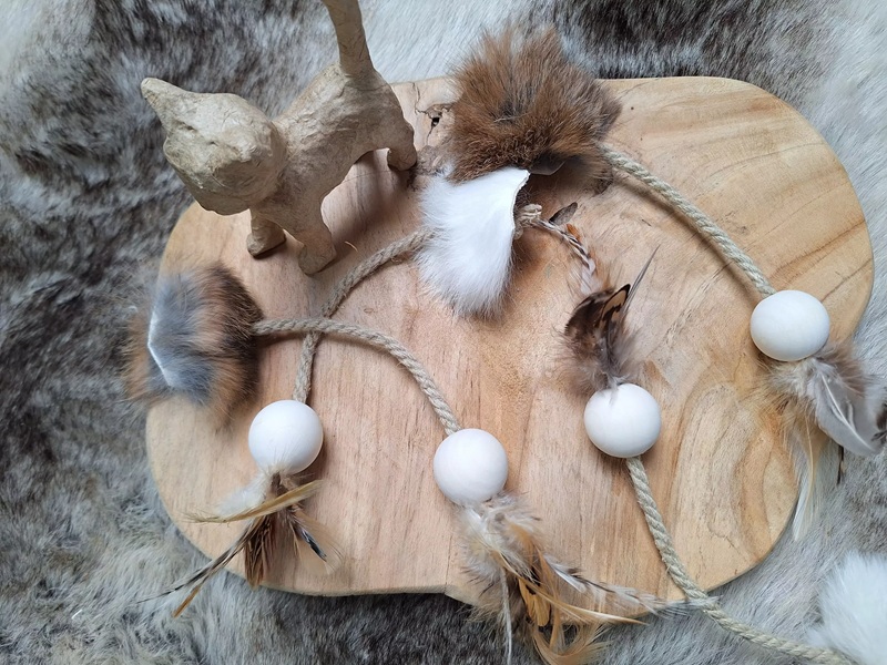 Holzbrett mit Spielzeug für Katzen: Papierkatze, Fellpuschel, Federn und Holzkugeln an Seilen.