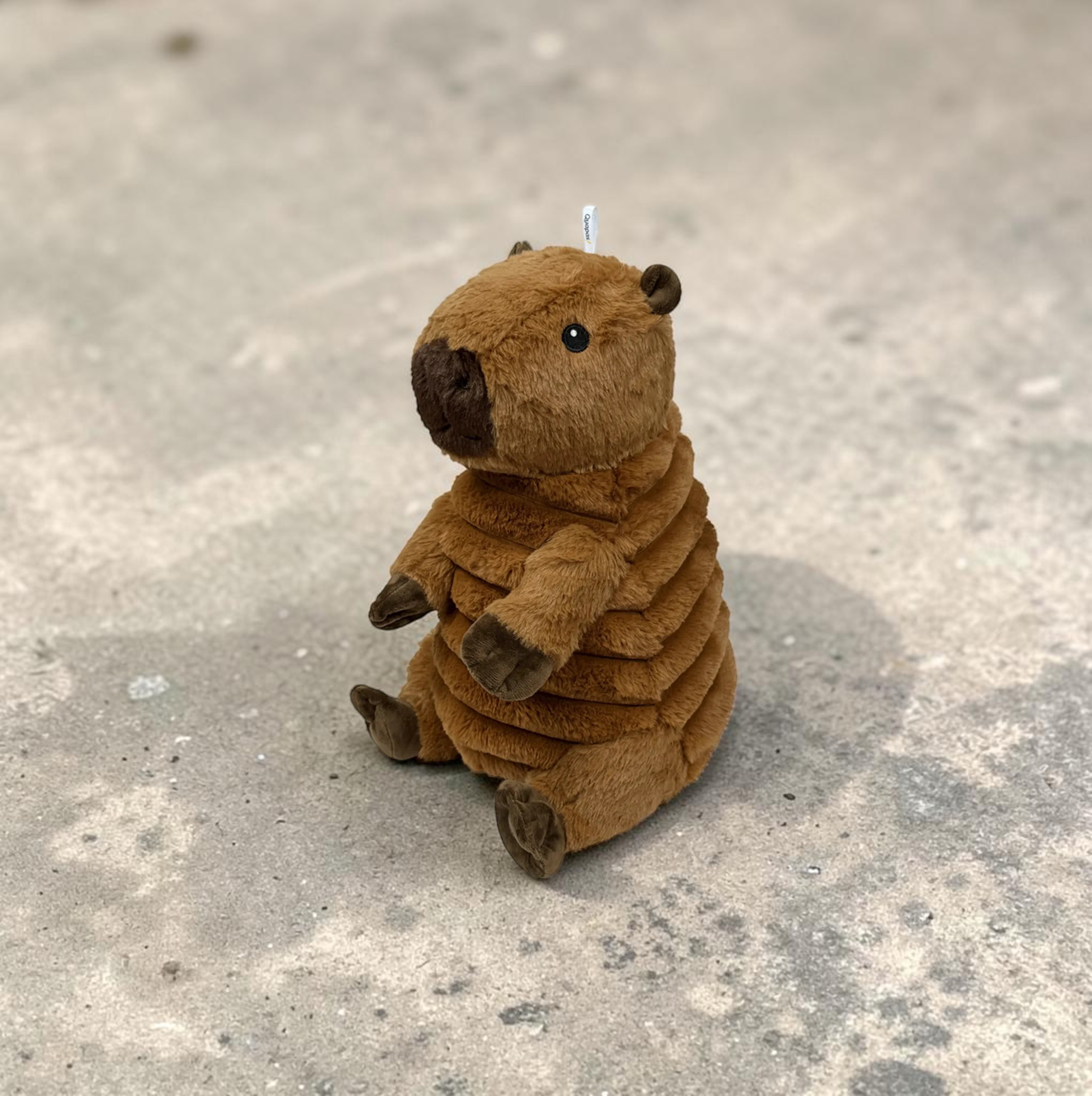 Ein sitzendes, braunes Plüschtier, das einem Capybara ähnelt, blickt leicht nach links. Es hat dunklere Pfoten, Ohren und Schnauze. Der Körper ist horizontal gerippt. Es sitzt auf hellgrauem, leicht verschmutztem Betonboden.