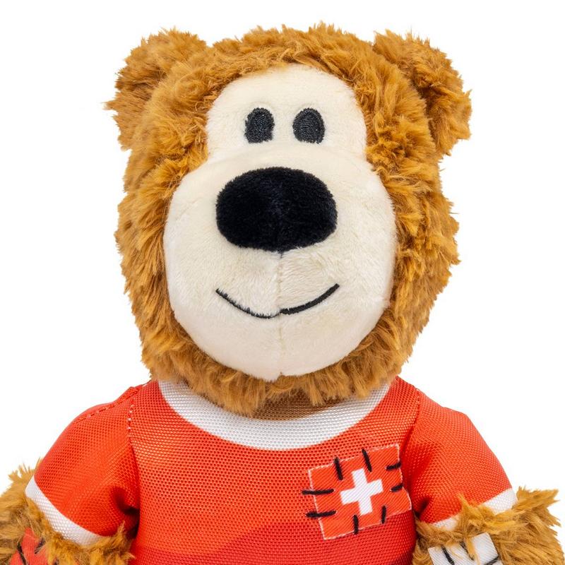 Un morbido orsacchiotto di peluche marrone guarda dritto nella fotocamera. Indossa una maglietta rossa con colletto bianco e la croce svizzera ricamata sul petto. Bocca chiara, naso nero, occhi neri e sorriso amichevole.