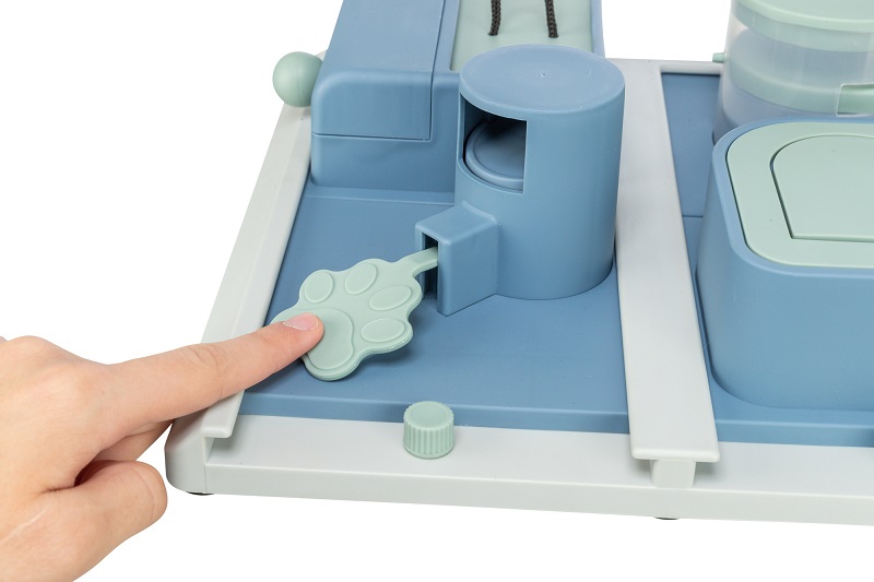 Un doigt appuie sur un bouton vert clair en forme de patte sur un jouet d’éveil bleu-gris pour animaux. Cela active un mécanisme cylindrique. À droite, des compartiments avec couvercles vert clair pour friandises.
