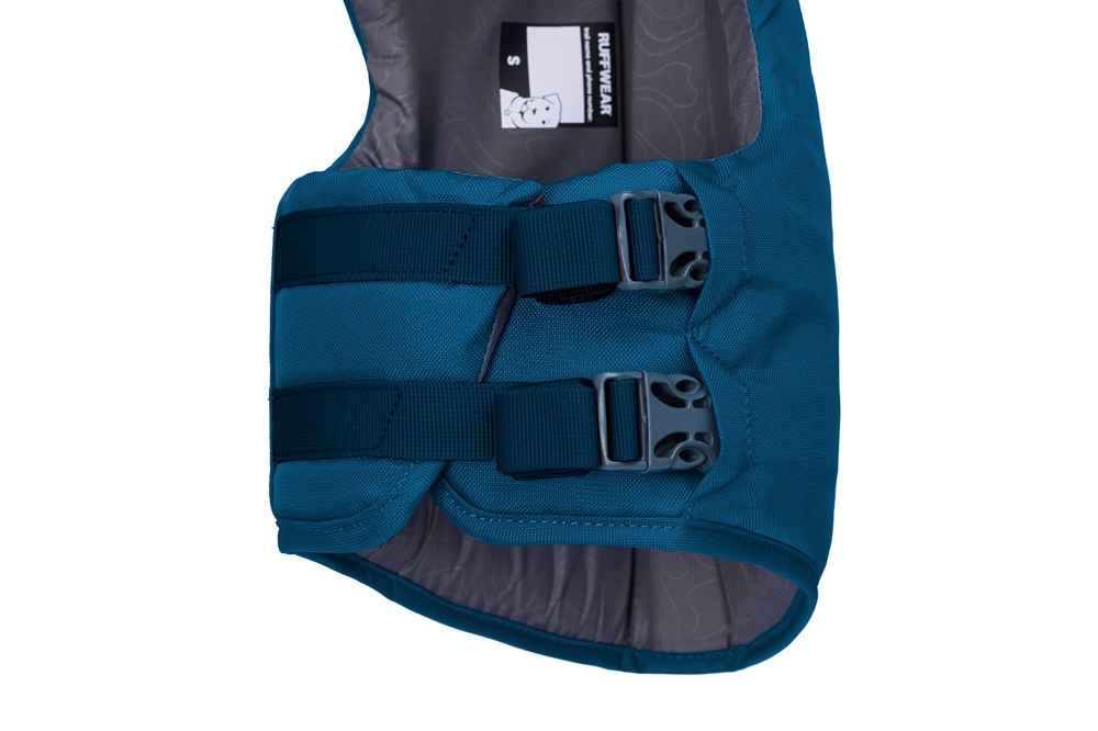 Gros plan sur le haut du dos d’un harnais pour chien Ruffwear bleu sarcelle. Deux sangles horizontales bleu foncé avec boucles en plastique grises sur tissu robuste. Doublure grise, étiquette Ruffwear, taille S.