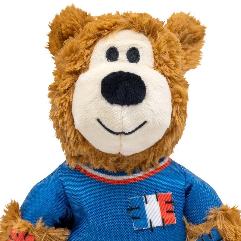 Orsacchiotto sportivo di peluche marrone con morbido pelo e muso simpatico. Muso bianco, naso di stoffa nero, piccoli occhi neri, bocca sorridente. Indossa una maglia blu con colletto rosso-bianco-blu e toppa con la bandiera francese.