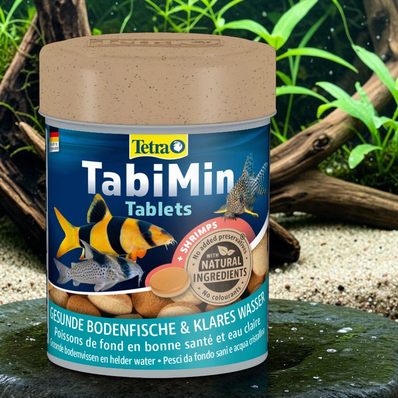 Blue container with a brown lid for Tetra ’s TabiMin fish food tablets. Label featuring three bottom-dwelling ornamental fish (yellow and black striped, spotted catfish) and brown tablets. In an aquarium with sand, driftwood and plants.
