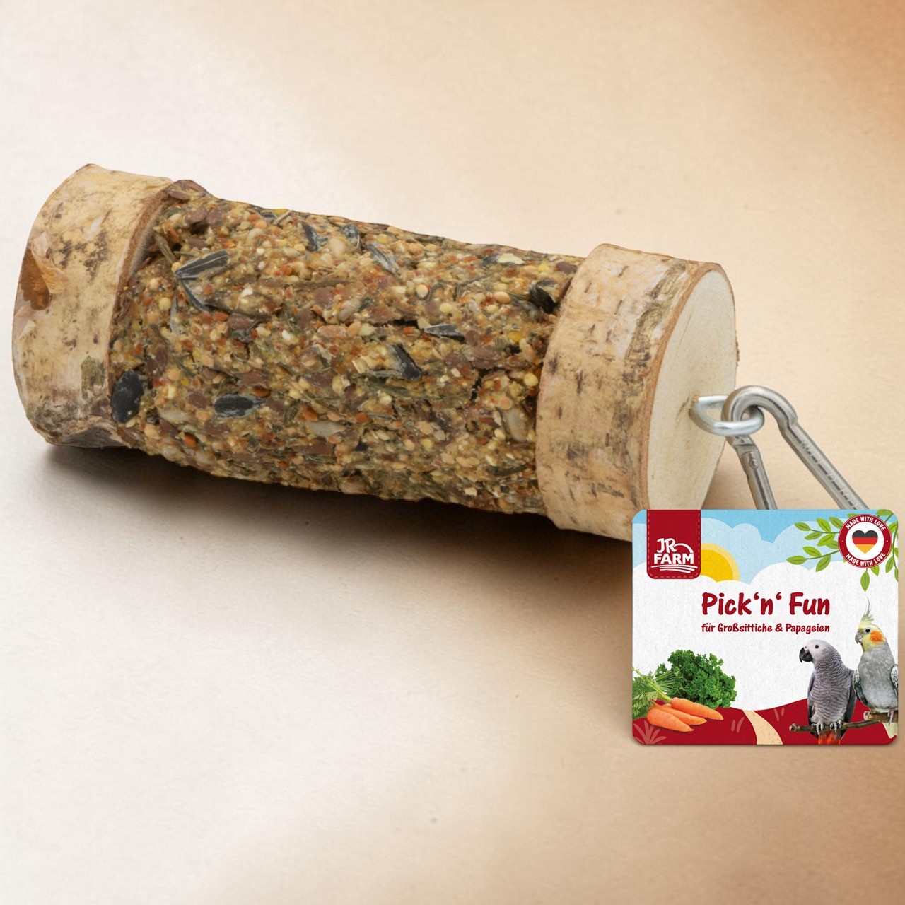 Cylindrical chew stick for birds with a seed/grain core, flanked on both sides by natural wooden discs. A silver metal hook on the right. Next to it is a square ‘Pick’n’ Fun’ label for parrots featuring two parakeets and vegetables.
