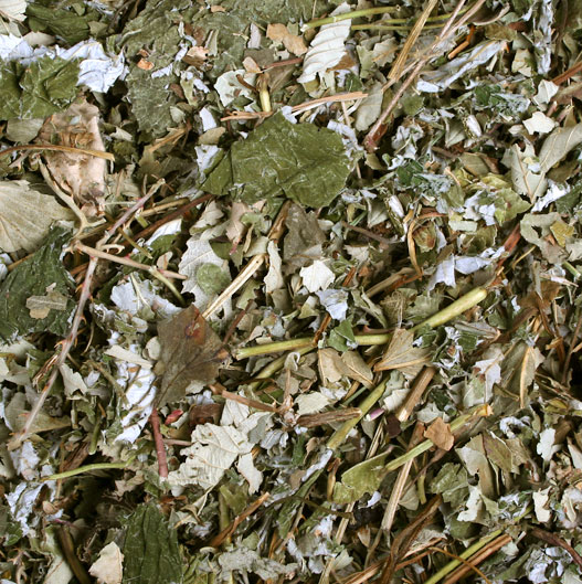 Nahaufnahme einer Mischung aus getrockneten Kräutern und Blättern in hellen bis dunklen Grüntönen, vermischt mit braunen und grauen Fragmenten. Kleine Stängel und zerkleinerte Blattstücke bilden eine raue, unregelmäßige Textur.