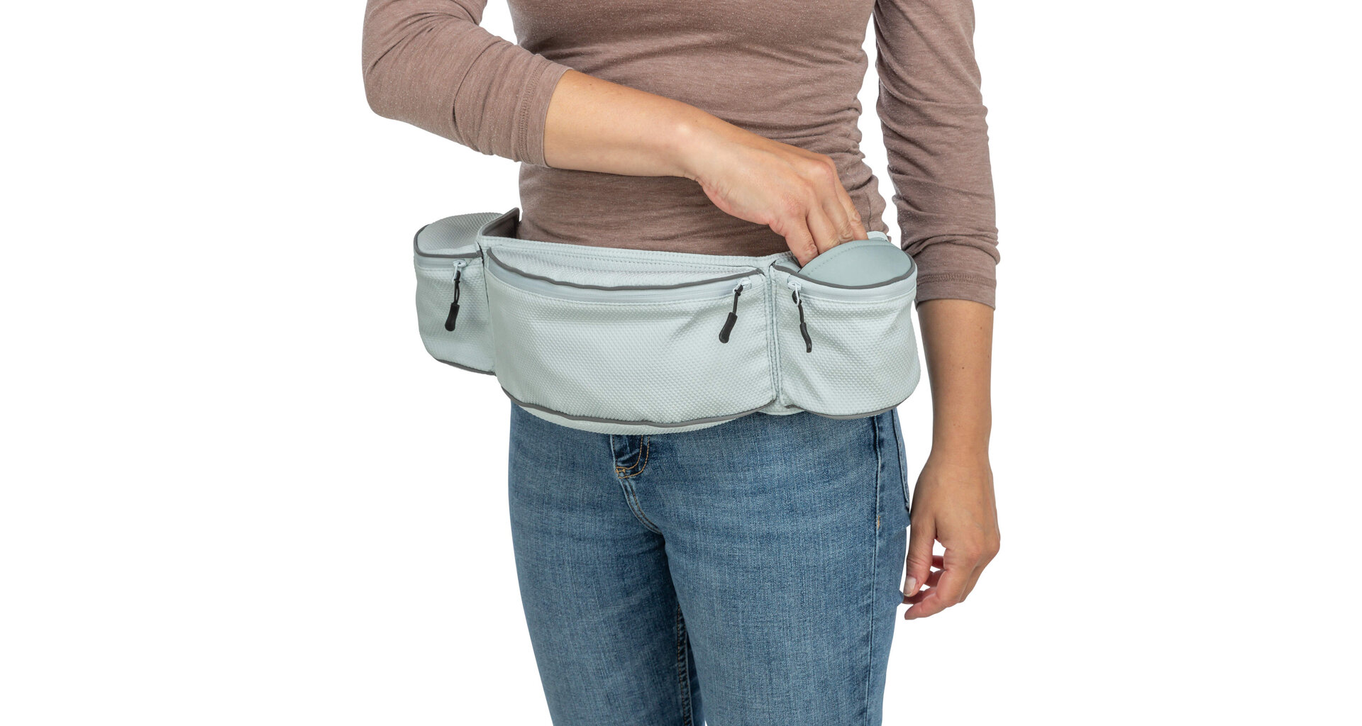 Eine Person trägt eine hellgraue Bauchtasche mit mehreren Reißverschlussfächern über blauen Jeans. Die rechte Hand greift in ein offenes Fach, um etwas herauszunehmen oder hineinzulegen. Strukturierte Oberfläche, graue Akzente.