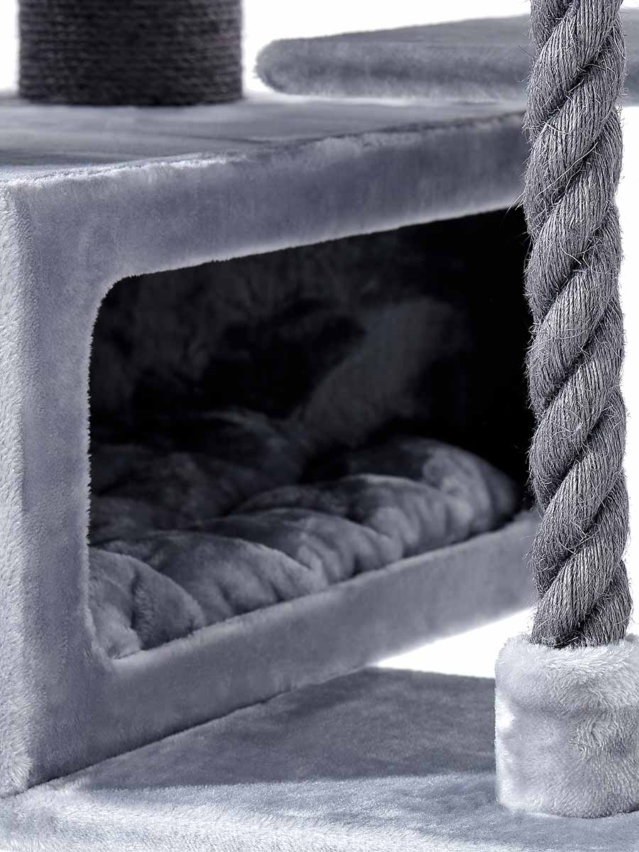 Close-up of a grey cat tree. A cosy cave with a dark, quilted cushion can be seen. To the right is a thick, twisted sisal scratching post. All surfaces are covered in soft, light grey plush fabric.