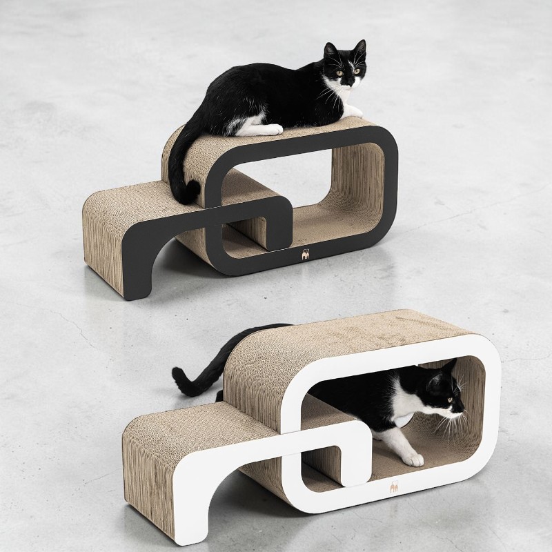 Un gatto bianco e nero è sdraiato su un tiragraffi sinuoso in cartone ondulato con cornice nera. Un altro gatto bianco e nero striscia attraverso il tunnel di un tiragraffi simile con cornice bianca. Entrambi offrono possibilità di gioco e di graffiare.