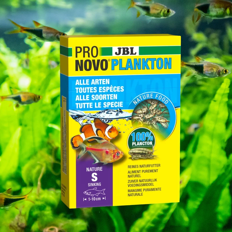 Gelbe JBL ProNovo Plankton S Packung vor grünem Aquarium, Fische schwimmen. Auf der Packung sind Clownfisch und ein Salmler abgebildet.