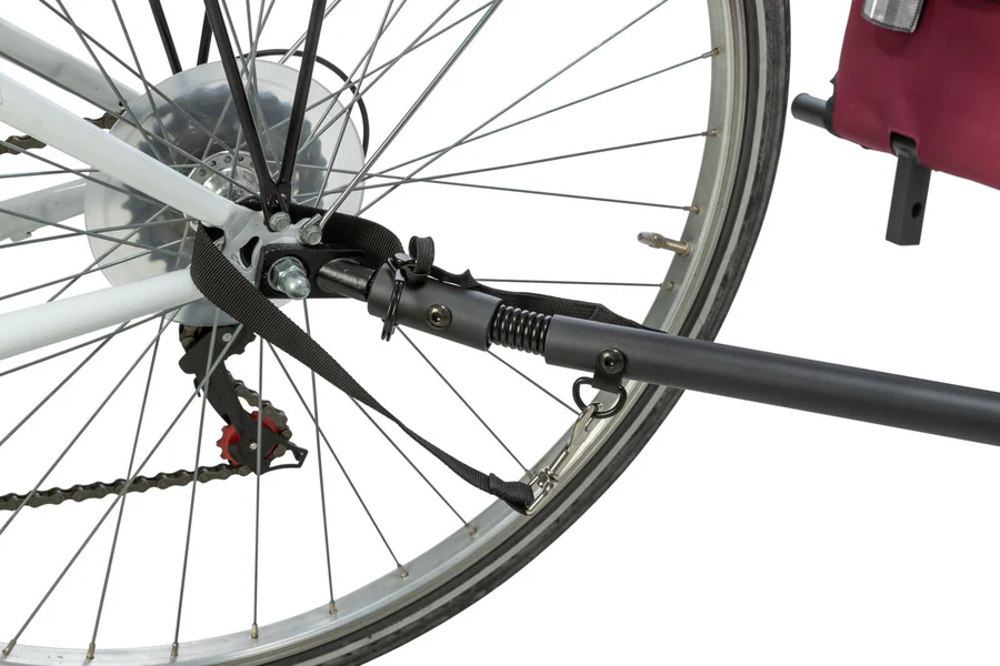 Nahaufnahme eines schwarzen Fahrradanhänger-Kupplungsmechanismus an der Hinterachse eines weißen Fahrrads. Ein schwarzes Zugrohr mit sichtbarer Feder ragt nach rechts. Schwarze Gurte sichern die Verbindung zum Hinterrad.