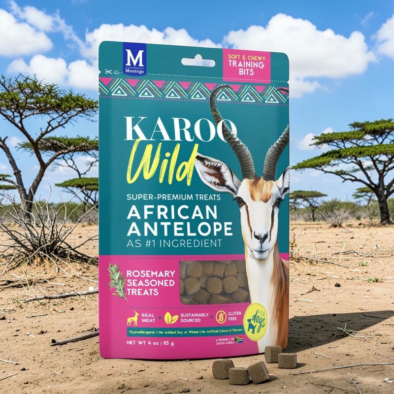 Una confezione di bocconcini per cani Karoo Wild con antilope africana e rosmarino è sulla sabbia asciutta. Sullo sfondo: acacie sotto un cielo blu con nuvole a pecorella. In primo piano, bocconcini quadrati.