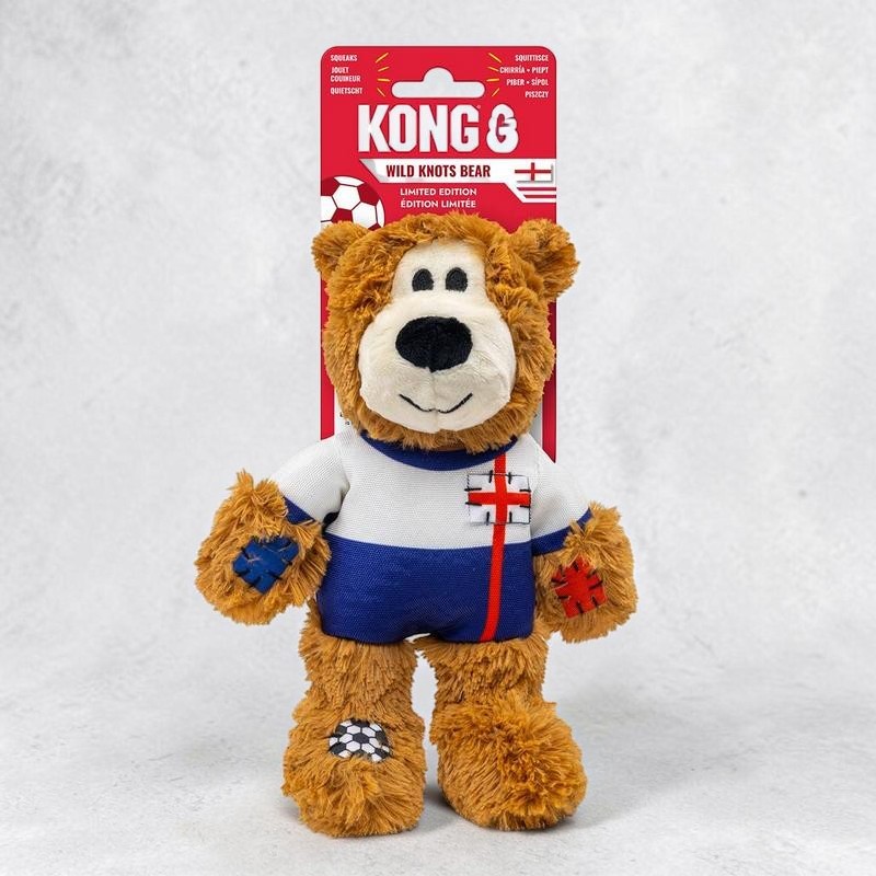 Orsacchiotto di peluche marrone con maglia da calcio bianca e blu e croce di San Giorgio rossa sul petto. Toppe a croce blu e rosse sulle zampe, pallone ricamato sul piede sinistro. Sfondo: confezione rossa Kong Wild Knots Bear.