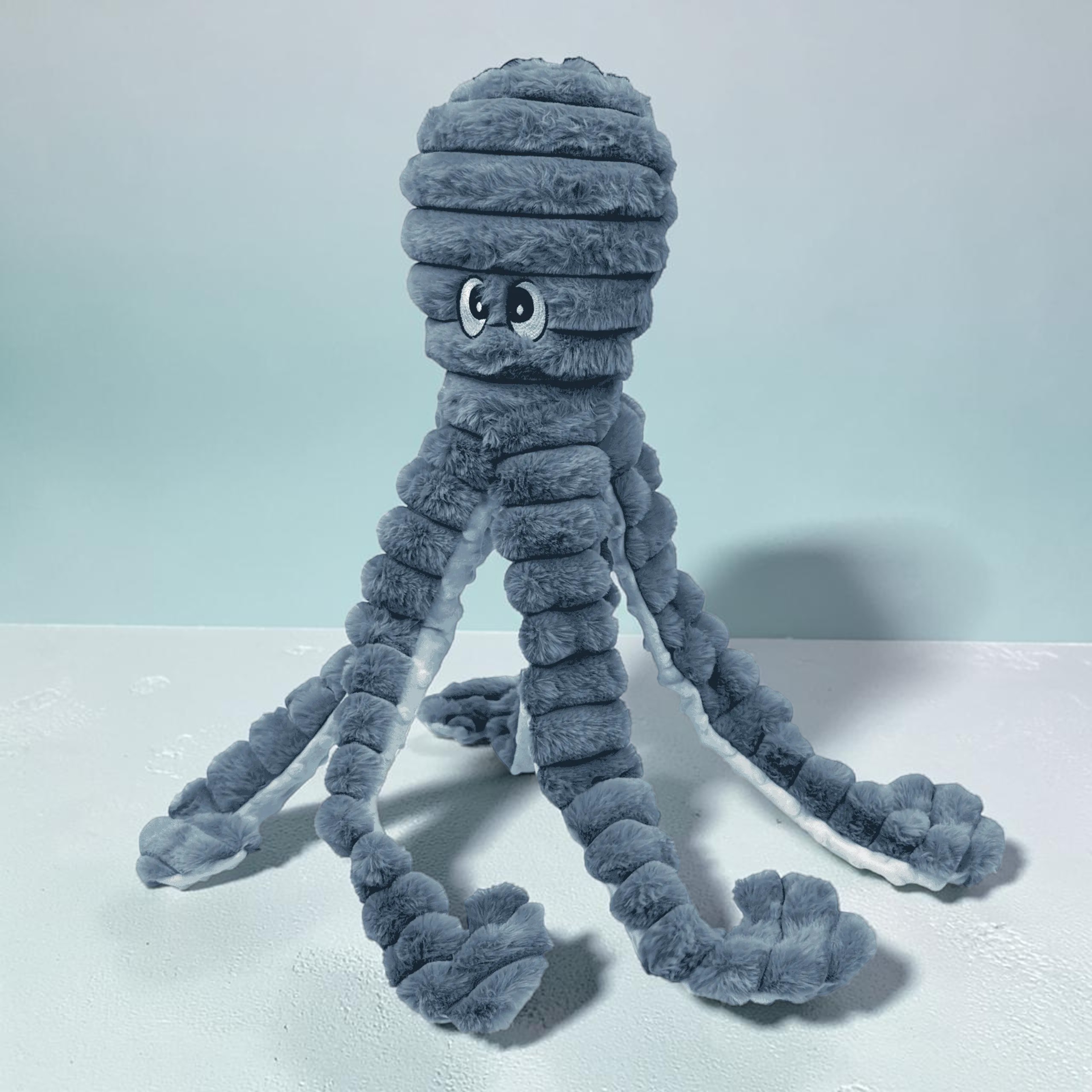 Großer, kuscheliger XXL-Oktopus aus grauem Plüsch auf hellem Untergrund vor dezent blauer Wand. Freundliche Augen, acht lange weiche Tentakel, gerippt und segmentiert, seitlich und nach vorn ausgebreitet.