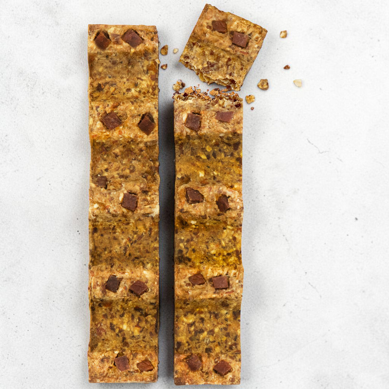 Due barrette allungate marrone dorato, con pezzi di noci e cioccolato fondente, su sfondo grigio chiaro. Quella a sinistra è integra; quella a destra è spezzata in alto, con un pezzo accanto e briciole.