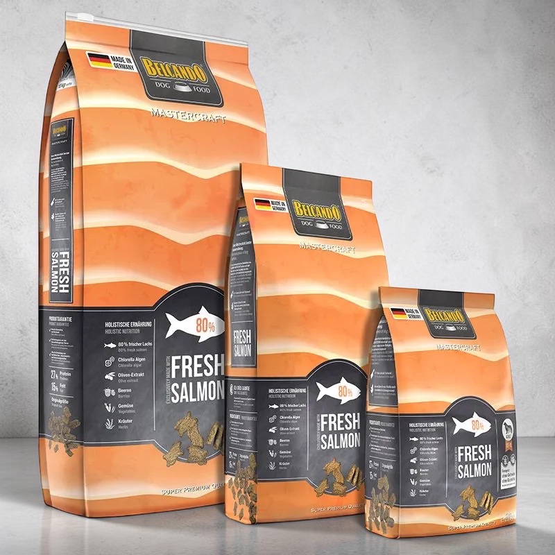 Drei orangefarbene Belcando Mastercraft "Fresh Salmon" Hundefutter-Säcke in verschiedenen Größen sind aufgereiht. Sie betonen "80% frischer Lachs" und holistische Ernährung. Knochenförmige Trockenfutter-Stücke liegen davor.