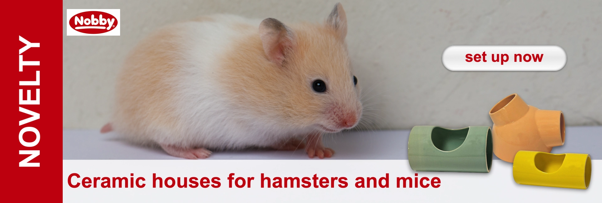 A small, fluffy hamster with light brown and white fur looks curiously to the right. Next to him are three colourful ceramic houses for small pets: a green one, a yellow tube-shaped house and an orange T-shaped house. For hamsters and mice.