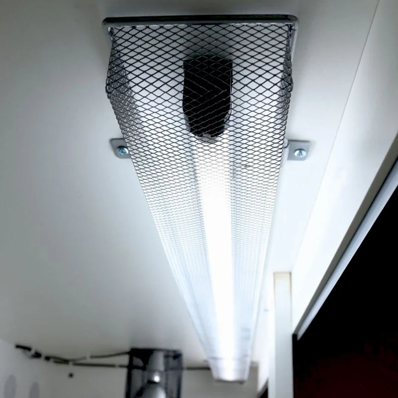 Ein helles, rechteckiges Lampen-Schutzgitter ist an der Unterseite einer weissen Decke befestigt. Das schwarze, rautenförmige Metallgitter umhüllt die Leuchte und schützt Terrarientiere vor Kontakt mit der heissen Lichtquelle.