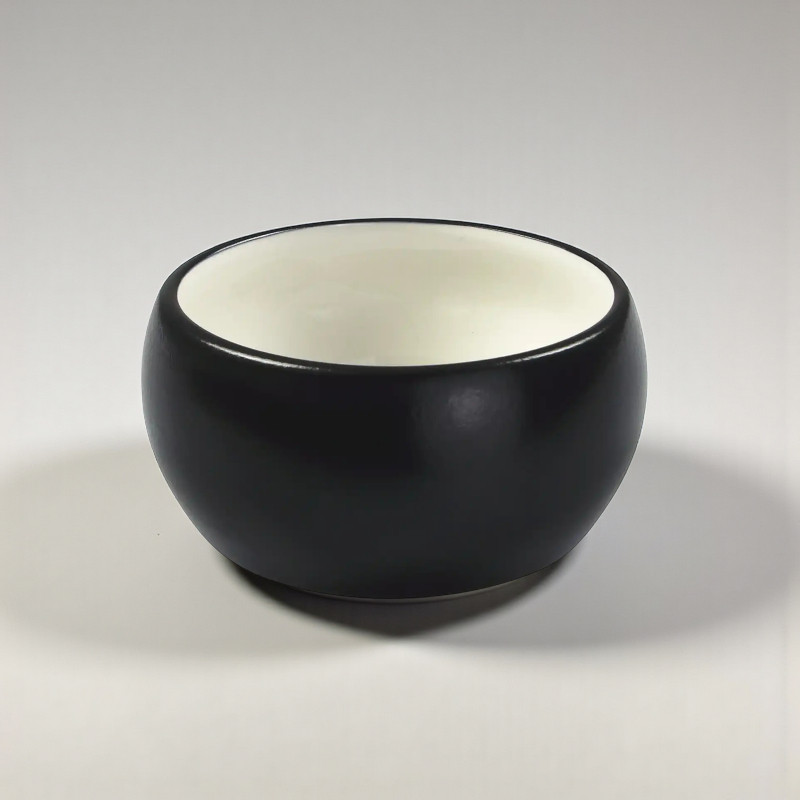 A round, black ceramic dog bowl with a white interior stands on a flat surface. The bowl is glossy and has a simple, modern shape. The inside of the bowl is smooth and reflects the light.