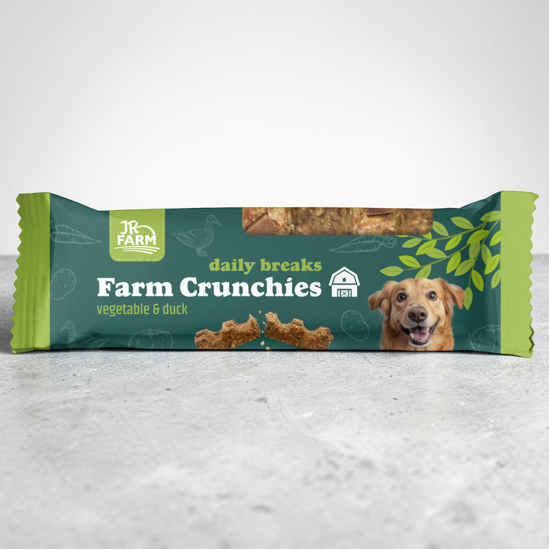 Green “JR Farm daily breaks” treat pack (Farm Crunchies vegetable & duck) on a grey background. Brown treats are visible; a broken piece lies in front. A smiling dog on the right.