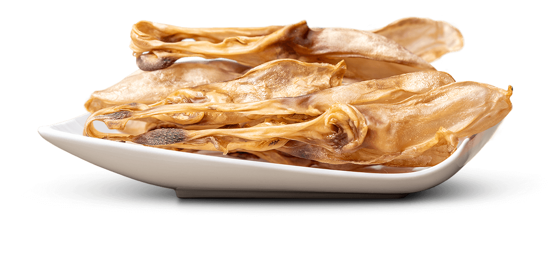 Goldbraune, getrocknete und gewellte Snacks, die an Ohren erinnern, liegen in einer weißen, flachen Schale. Einige Stücke haben dunklere, leicht gebräunte Ränder. Die unregelmäßigen Leckerlis sind locker gestapelt.