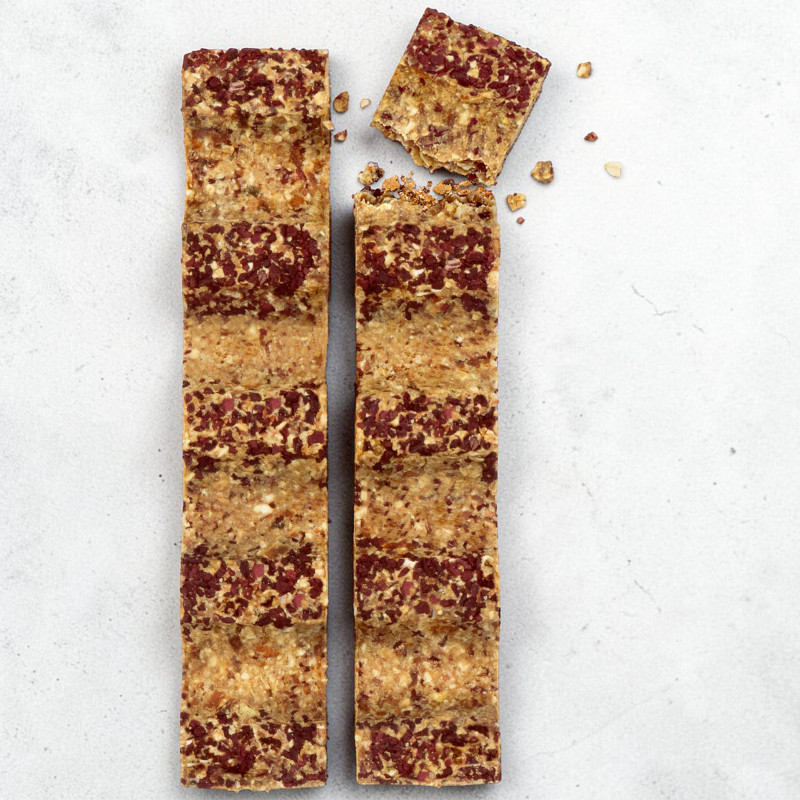 Due barrette di muesli verticali, base marrone chiaro con pezzetti di frutta o noci rosso scuro. La barretta a destra è spezzata in alto; accanto un piccolo pezzo quadrato, con briciole su sfondo grigio chiaro.
