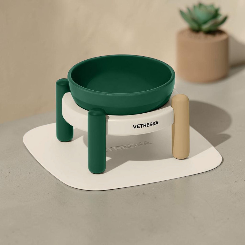 Ensemble de gamelles surélevé Vetreska avec une gamelle en céramique vert foncé sur socle blanc, deux pieds en bambou verts et un pied beige. Logo visible. Sur tapis silicone clair sur table grise, plante en pot floue.