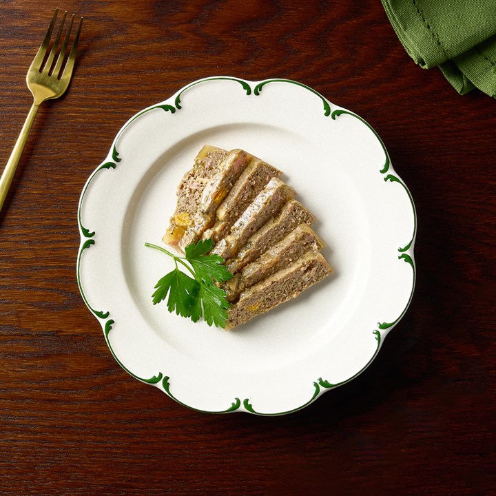 Un pâté allongé à base de poulet et de sanglier, coupé en tranches, est disposé de manière appétissante sur une assiette blanche incurvée au bord vert. Il est garni d'un brin de persil frais. Sa texture fine est bien visible.